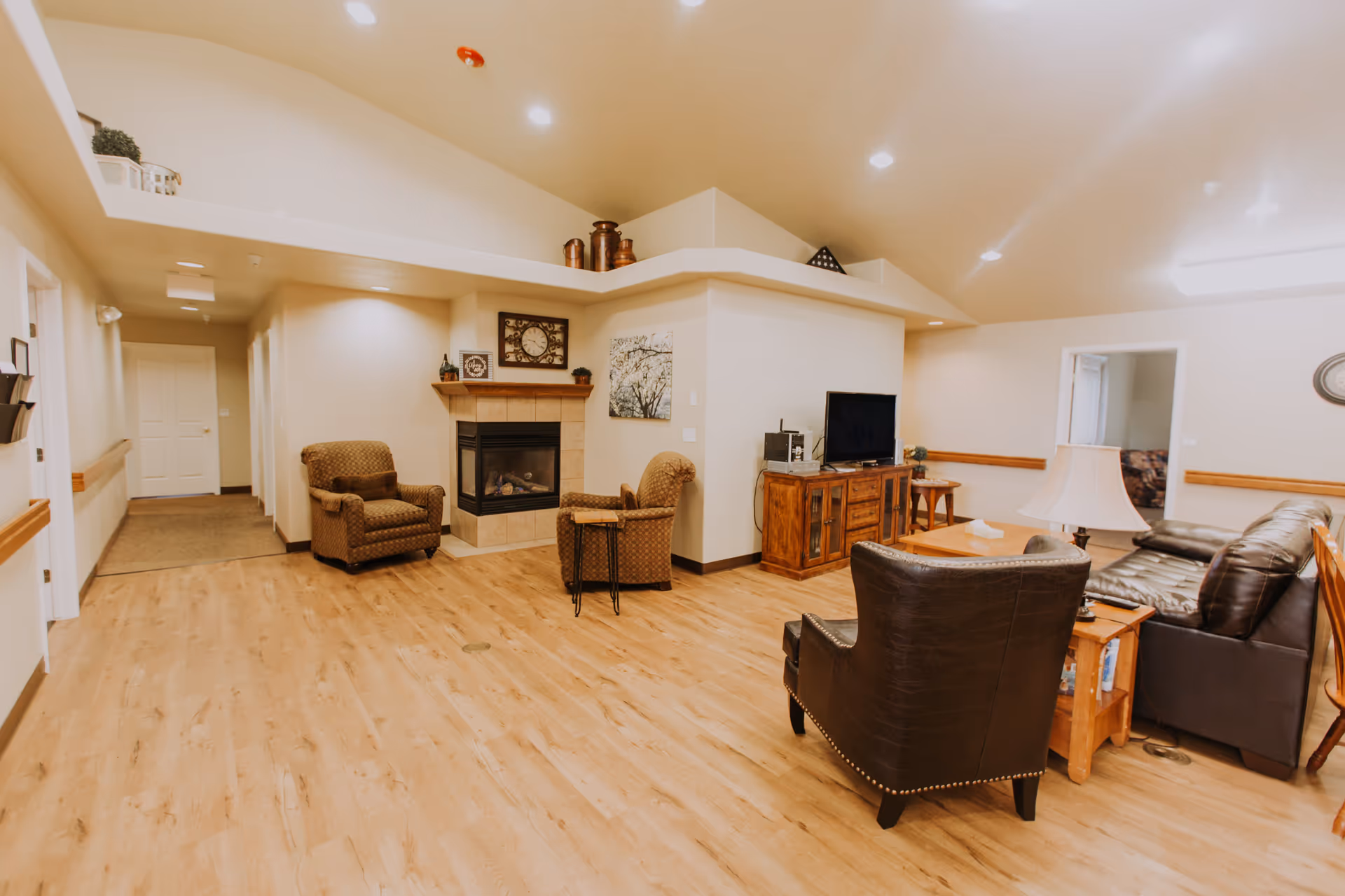 A cozy assisted living facility common area with wooden flooring, beige walls, and recessed lighting. The room features a corner fireplace with a clock and decorative items above it, two patterned armchairs, a dark leather armchair, a leather sofa, a wooden coffee table, a wooden TV stand with a flat-screen TV, and a table lamp. There is a hallway with doors leading to other rooms and a small side table with a plant.