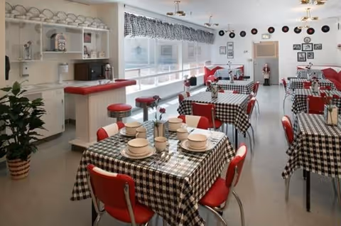 A bright dining room with multiple tables covered in black and white checkered tablecloths, each set with plates, cups, and utensils. Red chairs surround the tables, and there are red cushioned stools at a counter with white cabinetry and shelves holding dishes and a microwave. The room has large windows with checkered valances, decorative plates on the wall, and framed pictures. A potted plant is visible in the corner.