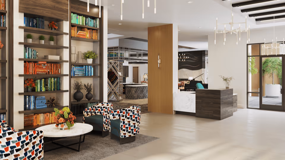 A modern and spacious assisted living facility common area with colorful patterned armchairs around a round white marble coffee table with a flower arrangement. There are tall bookshelves filled with books and decorative plants on the left side. The reception desk is visible in the background along with large windows and doors leading to an outdoor area with greenery.