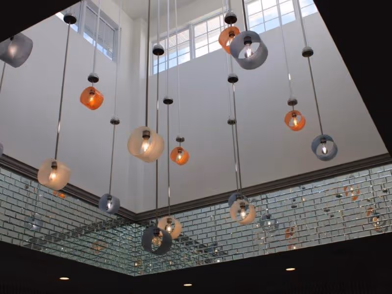 Ceiling view of a modern interior space featuring multiple hanging pendant lights with colorful, translucent shades in orange, blue, and white. The ceiling is high with large windows letting in natural light, and the walls below the ceiling are lined with reflective glass bricks.