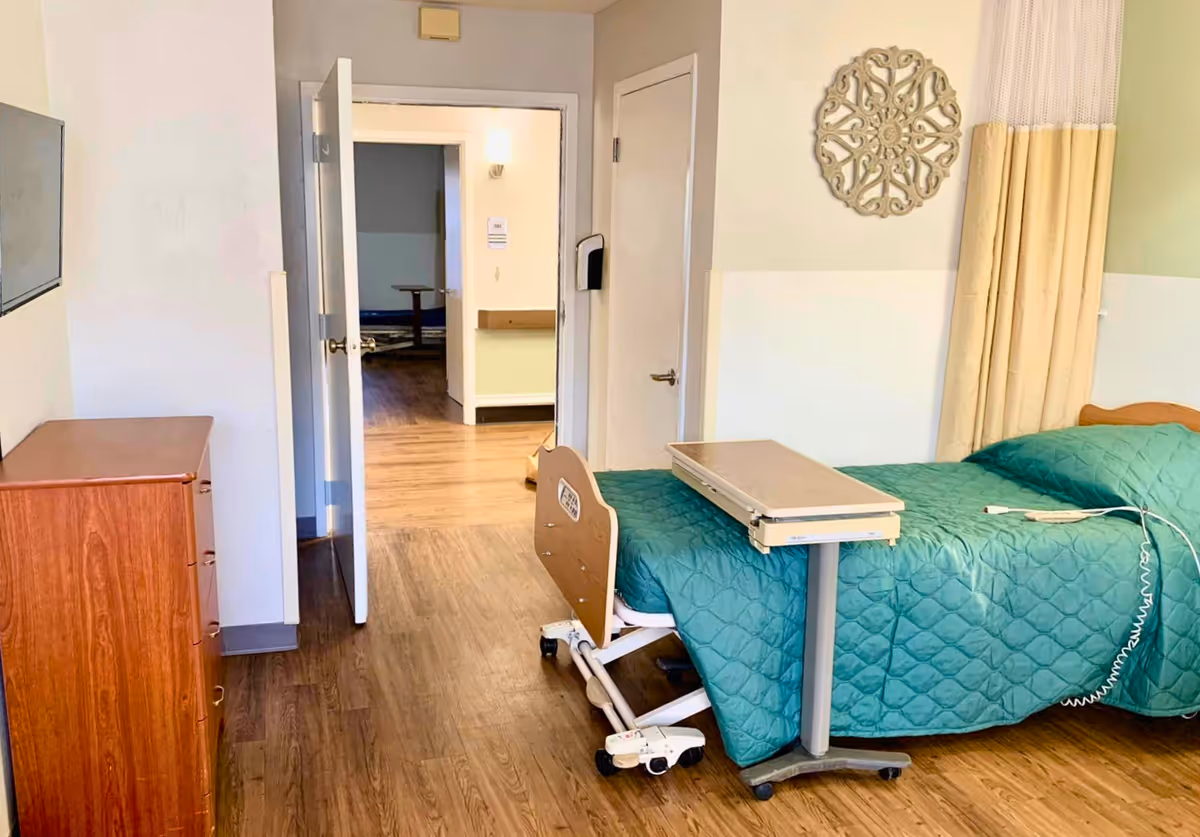 A senior care bedroom with a teal-covered adjustable bed, overbed table, wooden dresser, and an open door to a hallway.