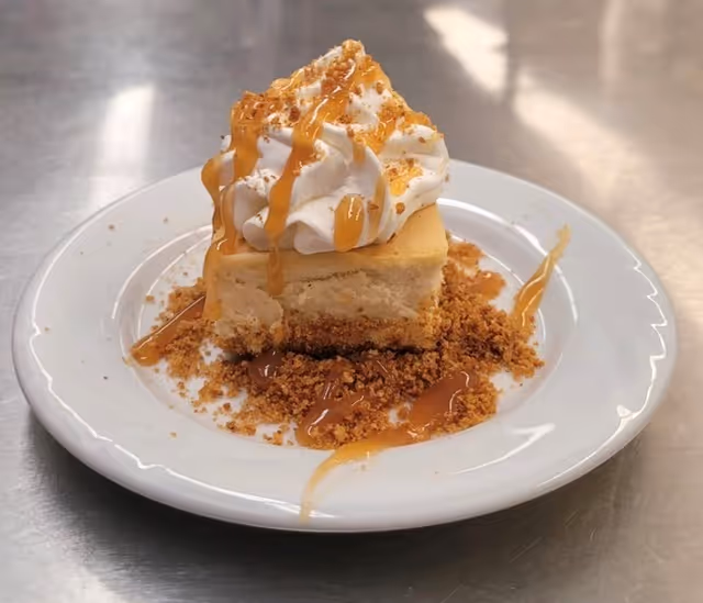 A plated slice of cheesecake topped with whipped cream and caramel drizzle on a white plate.
