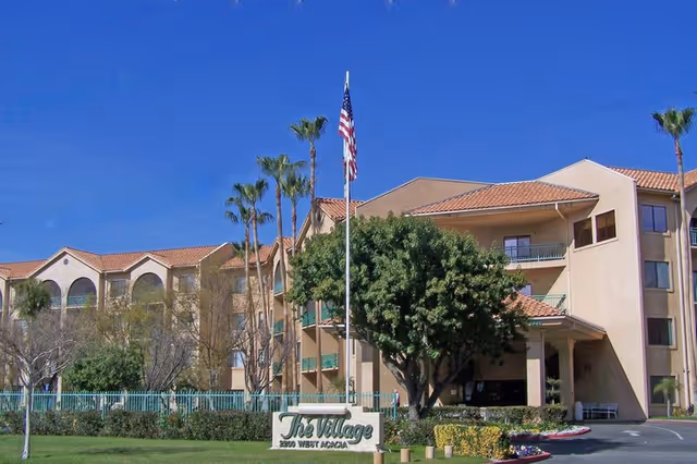 Front exterior of The Village senior living building with a sign, palm trees, and an American flag.