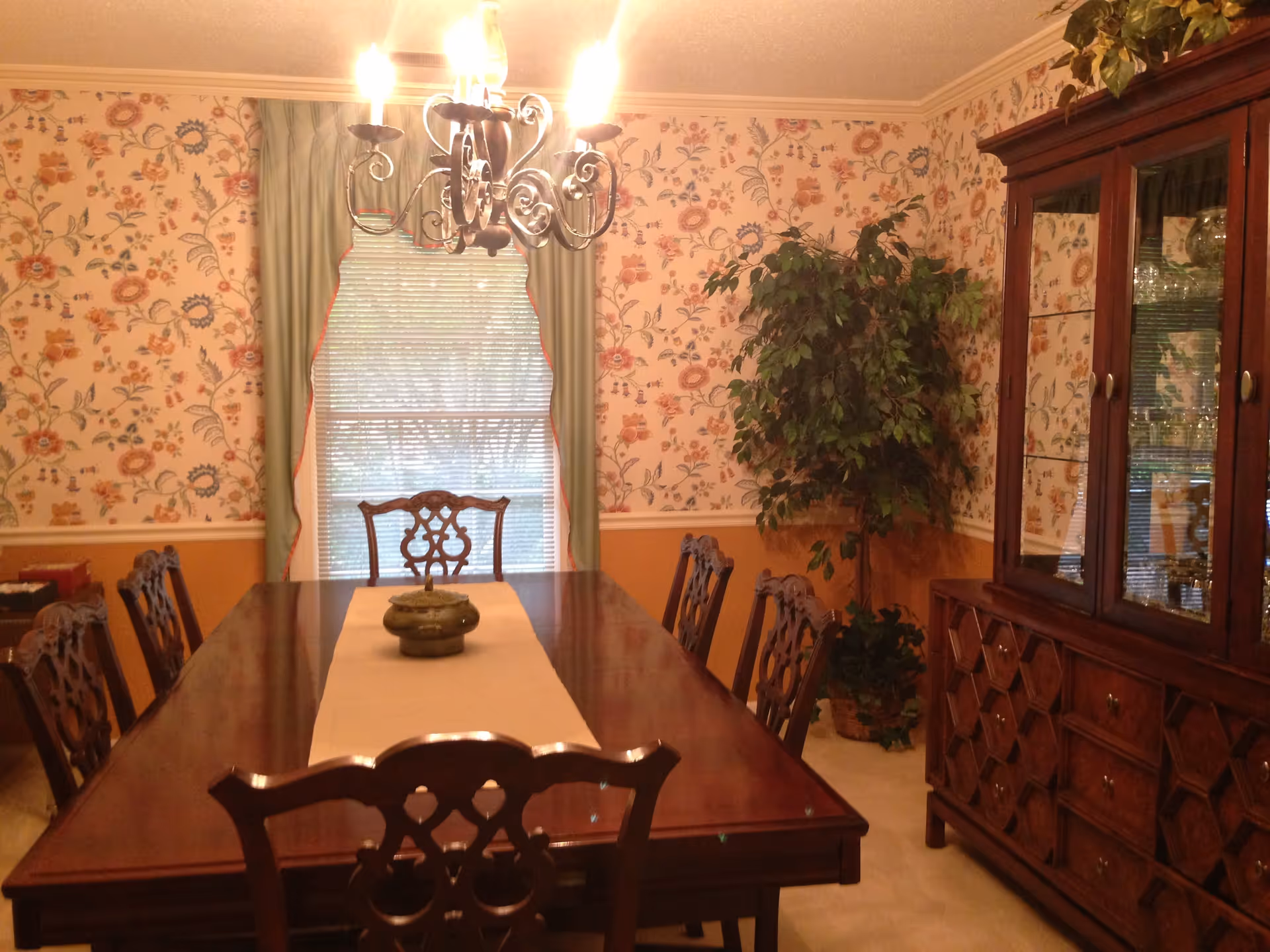 Formal dining room with a long wooden table and chairs, chandelier, floral wallpaper, a china cabinet, and a potted plant.