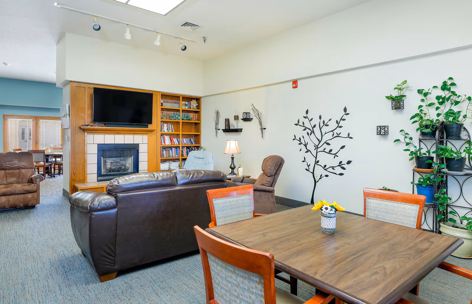 A cozy living room area in an assisted living facility featuring a brown leather couch, two recliner chairs, a wooden bookshelf with books, a flat-screen TV mounted above a fireplace, a wooden table with four chairs, and several potted plants along with a decorative wall decal of a tree.