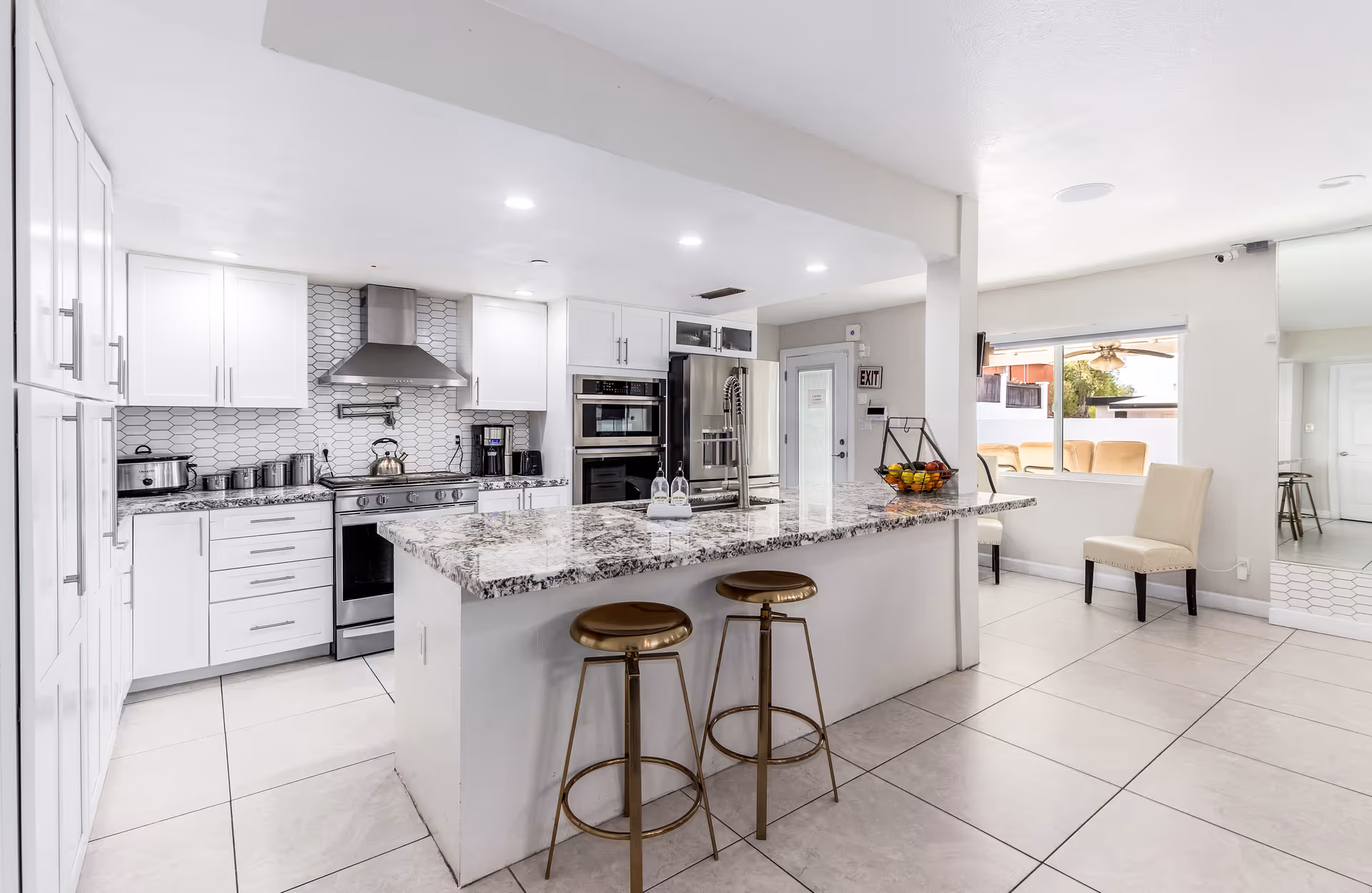 Bright modern kitchen with white cabinets, a large granite island with two bronze bar stools, stainless steel appliances including a stove, oven, microwave, and refrigerator. There is a window showing an outdoor seating area and a door with an exit sign above it.