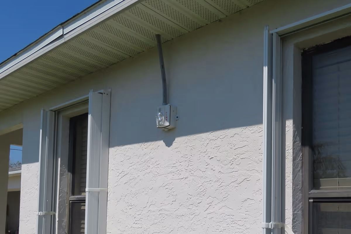 Exterior wall of a building with a window and white shutters, a mounted light fixture, and a textured stucco surface under a clear blue sky.