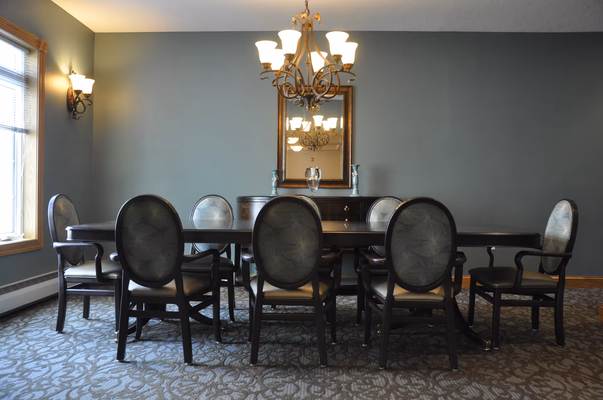 A dining room with a long dark wooden table surrounded by eight upholstered chairs. A chandelier with multiple lights hangs above the table, and a large mirror is mounted on the wall behind a sideboard. The room has a window with blinds on the left and patterned carpet flooring.