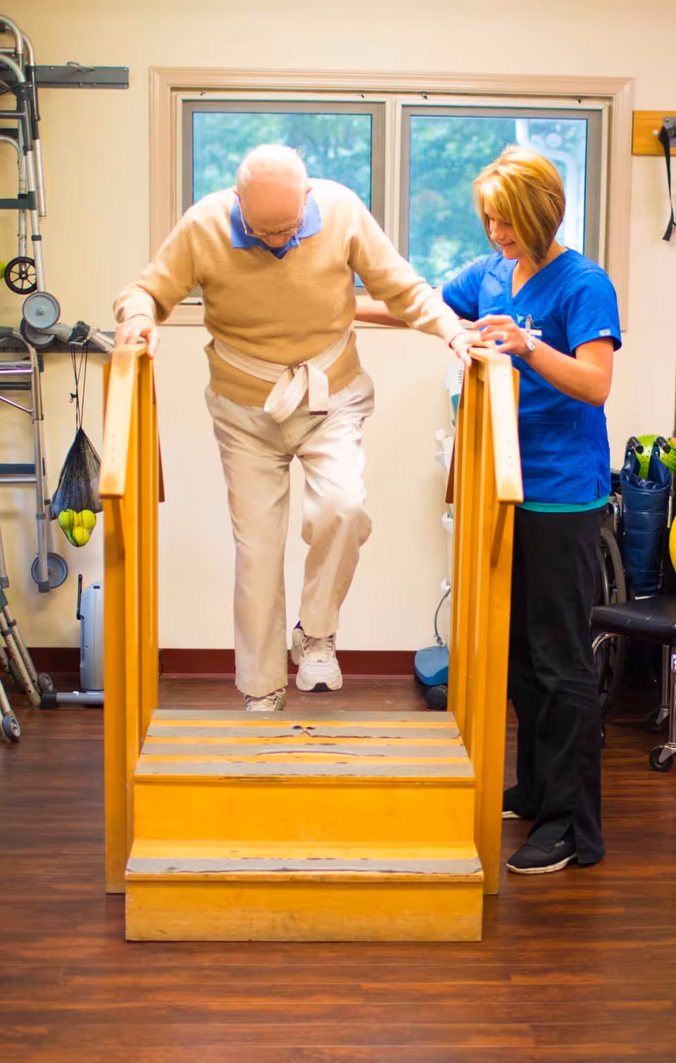 An elderly man wearing a beige sweater and light pants is walking up wooden rehabilitation stairs while holding the handrails. A female healthcare worker in blue scrubs is standing beside him, providing support and encouragement. The room has a window in the background and various medical equipment visible on the sides.