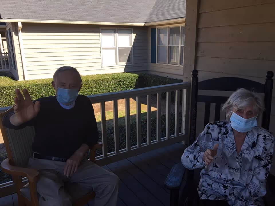 Two elderly individuals wearing face masks sitting on a covered porch with wooden railing. One person is waving while the other is gesturing with their hand. Behind them is a building with beige siding and windows, along with neatly trimmed bushes.