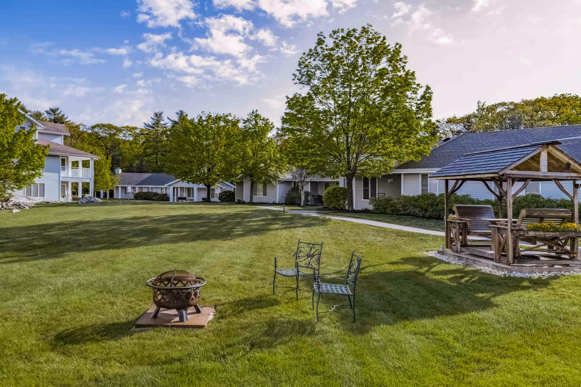 Well-maintained grassy courtyard with a fire pit, metal chairs, a wooden gazebo swing, trees, and surrounding senior living buildings under a partly cloudy sky.