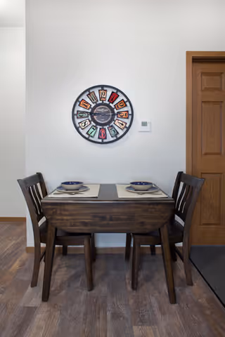 Small wooden dining table set for two against a white wall with a decorative round clock and a wooden door nearby.