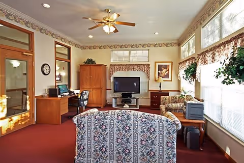 A cozy living room area with floral patterned sofas, a large flat-screen TV on a stand, wooden furniture including a desk with a computer, and windows with floral valances letting in natural light. The room has a ceiling fan with lights and a red carpeted floor.