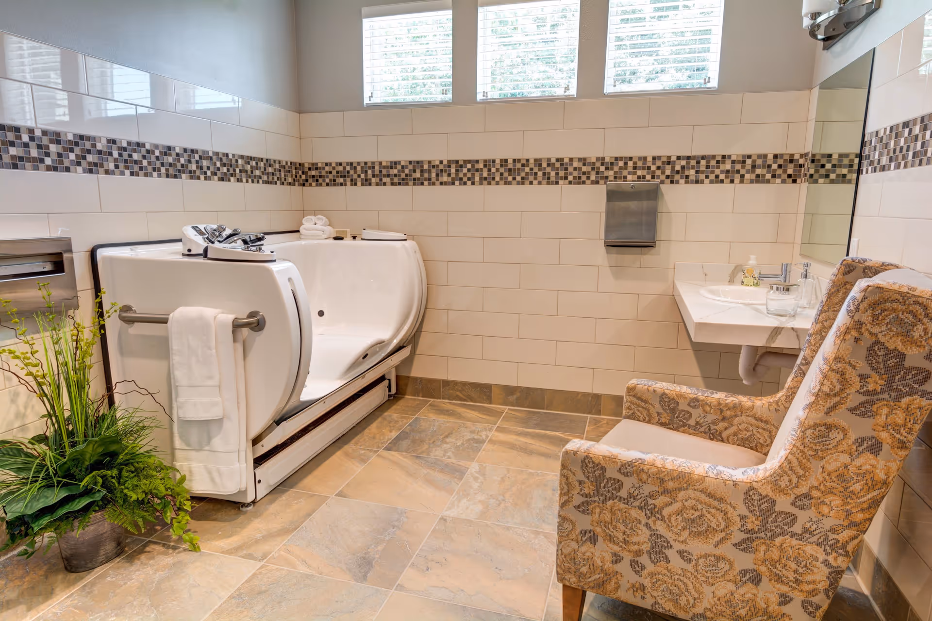 A spacious bathroom with beige tiled floor and white tiled walls featuring a decorative mosaic border. There is a walk-in bathtub with a towel hanging on its side, a floral patterned armchair, a wall-mounted sink with soap and a glass, a stainless steel paper towel dispenser, and a green potted plant in the corner. Three windows near the ceiling allow natural light into the room.