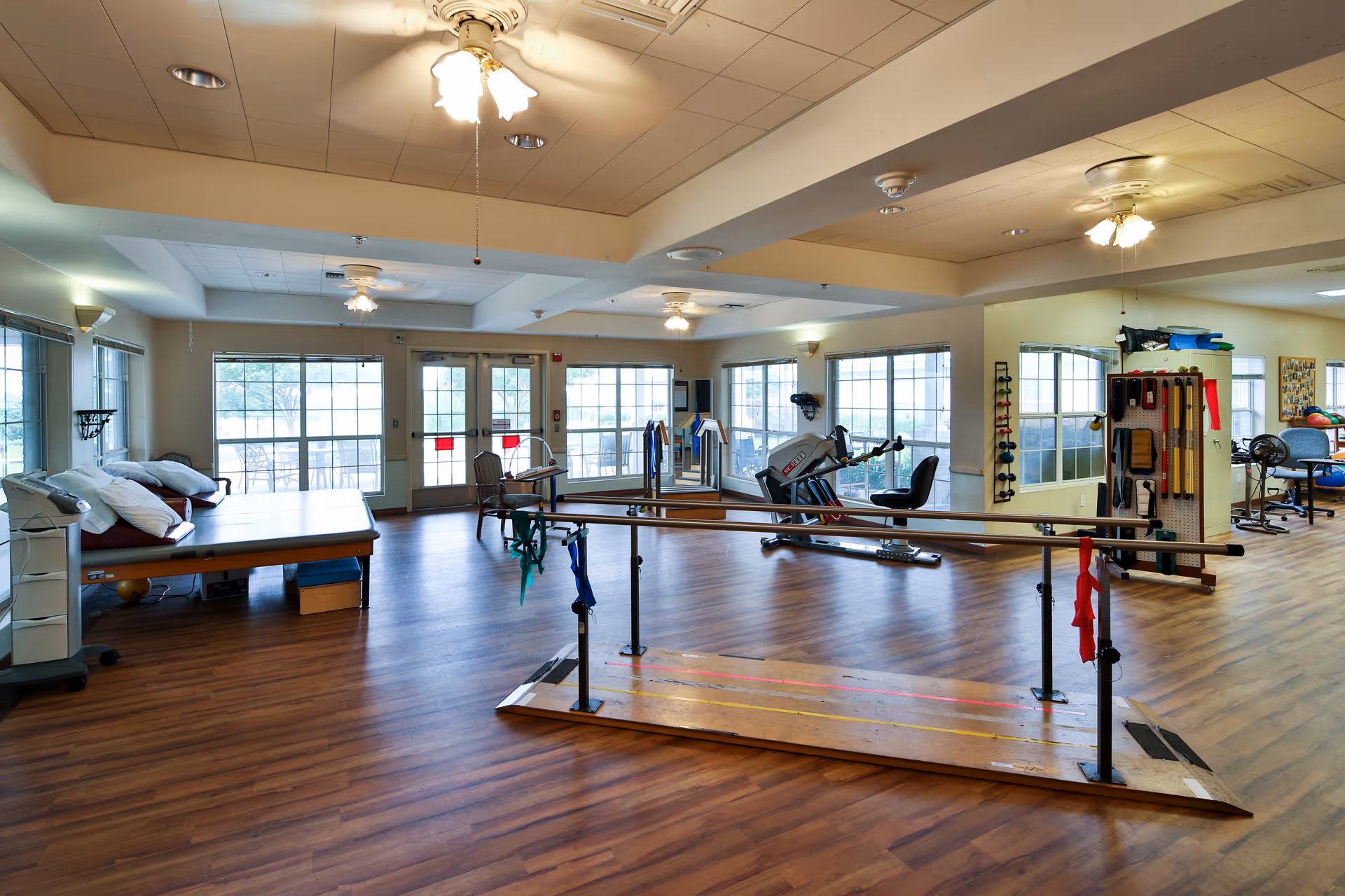 A spacious rehabilitation room with wooden flooring, parallel bars for walking exercises, exercise equipment, therapy tables with pillows, and large windows letting in natural light.