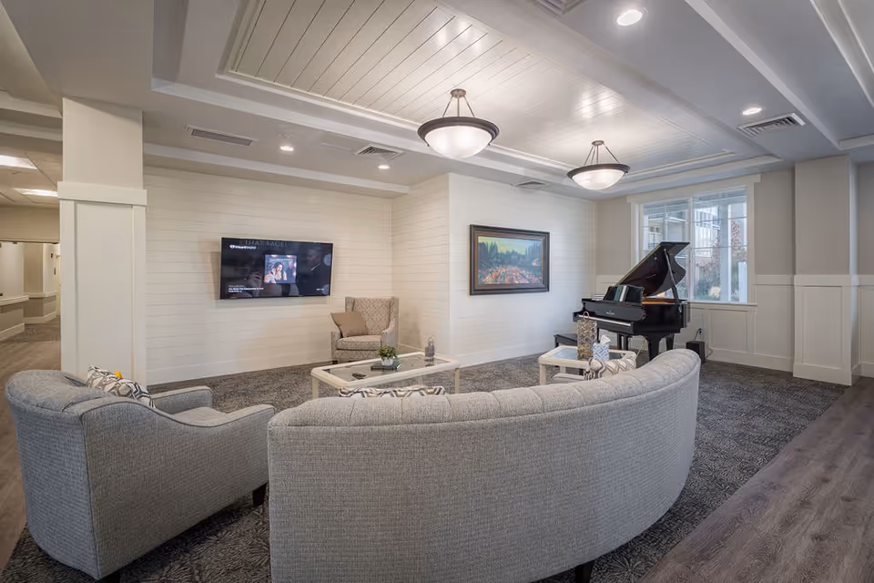 A cozy senior living common area with a curved gray sofa, an armchair, a coffee table, a wall-mounted flat screen TV, a framed landscape painting, and a grand piano near a window. The room features recessed lighting and a white paneled ceiling.