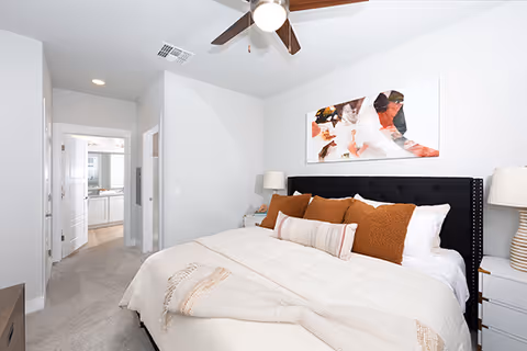 A neatly made bed with white and brown pillows and a beige throw blanket in a bedroom. The room has light gray walls, a ceiling fan with a light, a bedside table with a lamp, and an abstract painting above the bed. A hallway leading to a bathroom and other rooms is visible in the background.