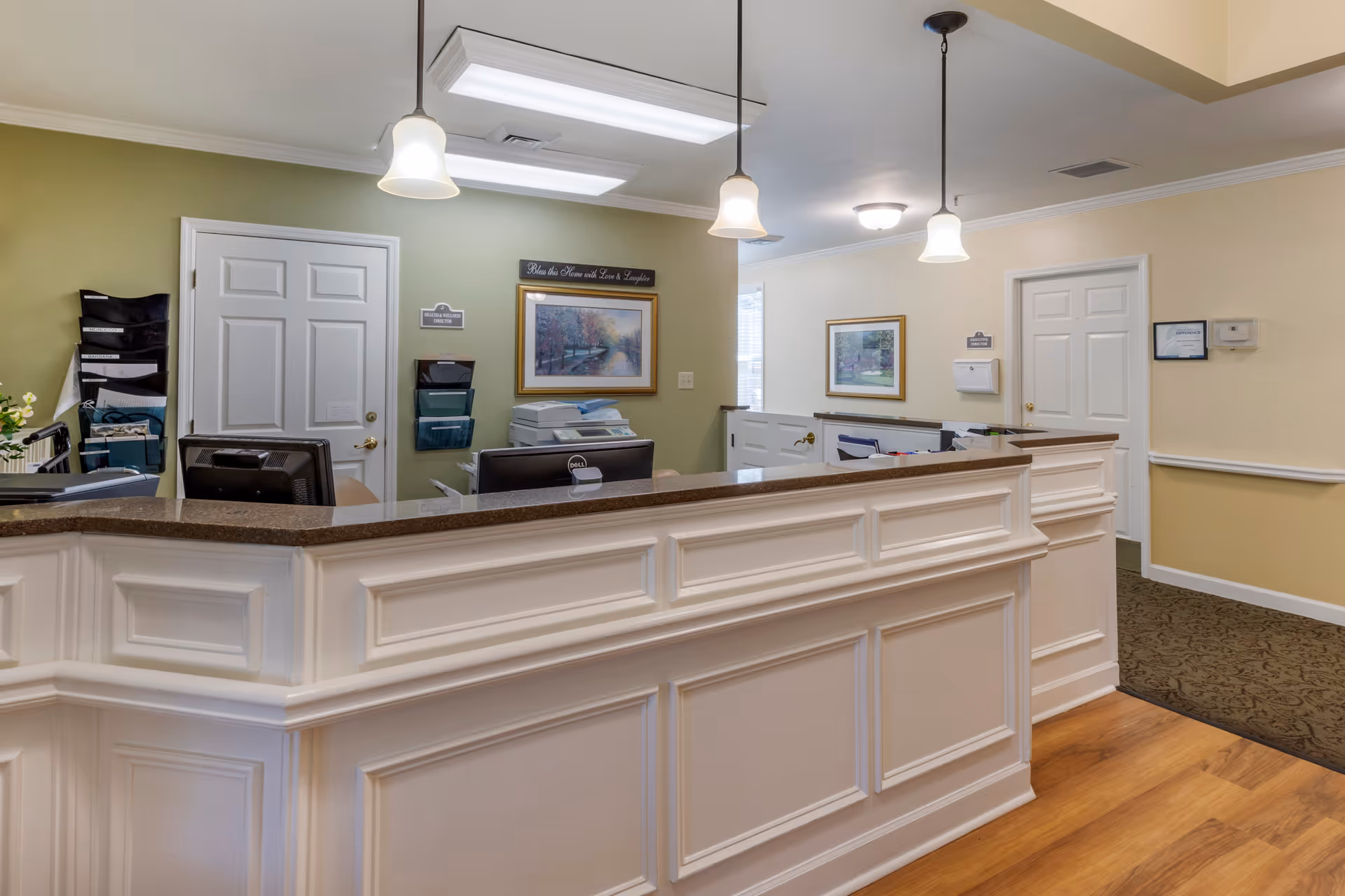 Reception desk area with a white paneled counter, pendant lights, computers, and doors in a warmly painted interior.