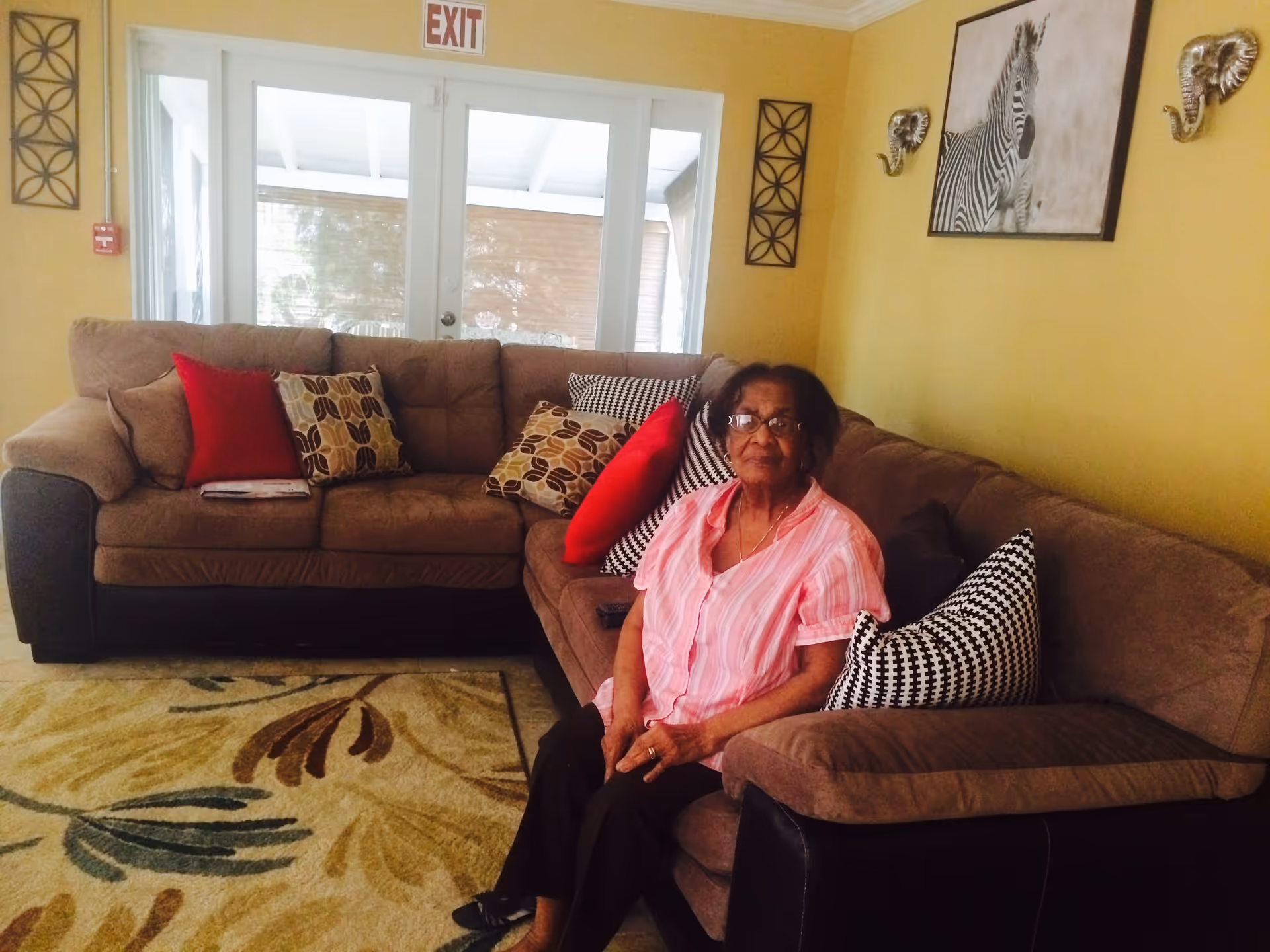 An elderly woman wearing glasses and a pink striped shirt is sitting on a brown sectional sofa in a living room with yellow walls. The sofa has various patterned cushions in red, brown, and black and white. Behind the sofa, there are glass doors with an exit sign above them. The walls are decorated with a zebra painting and two metallic elephant head sculptures. A patterned rug covers the floor.