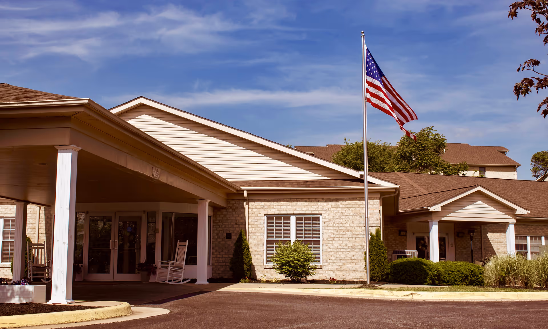 Exterior view of The Maynard senior living facility showing a single-story building with beige brick walls, white trim, and a covered entrance with rocking chairs. An American flag is flying on a flagpole in front of the building under a partly cloudy blue sky.