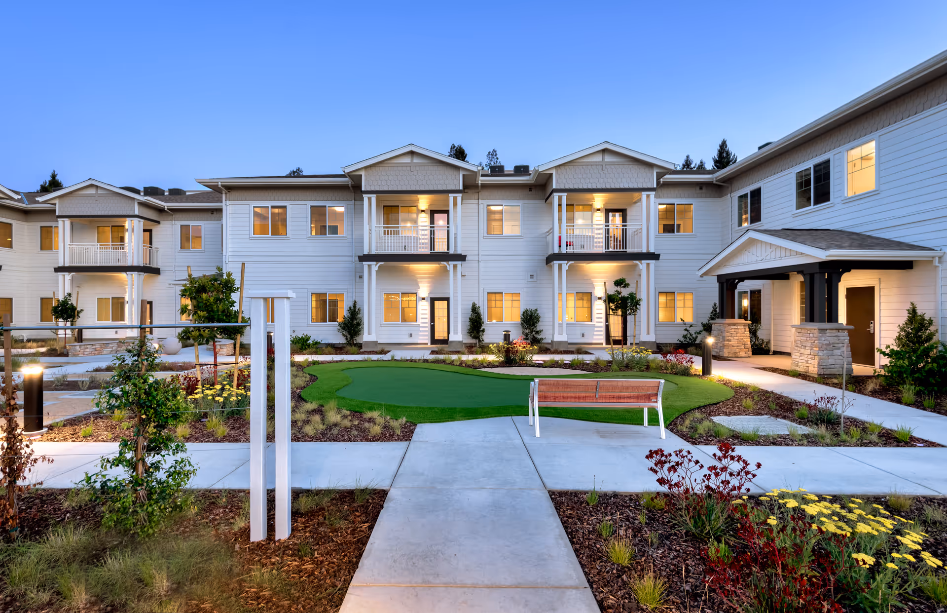 Exterior view of a two-story senior living facility building at dusk with well-lit windows and balconies. The foreground features a landscaped garden with a small putting green, a wooden bench, paved walkways, and various plants and flowers.