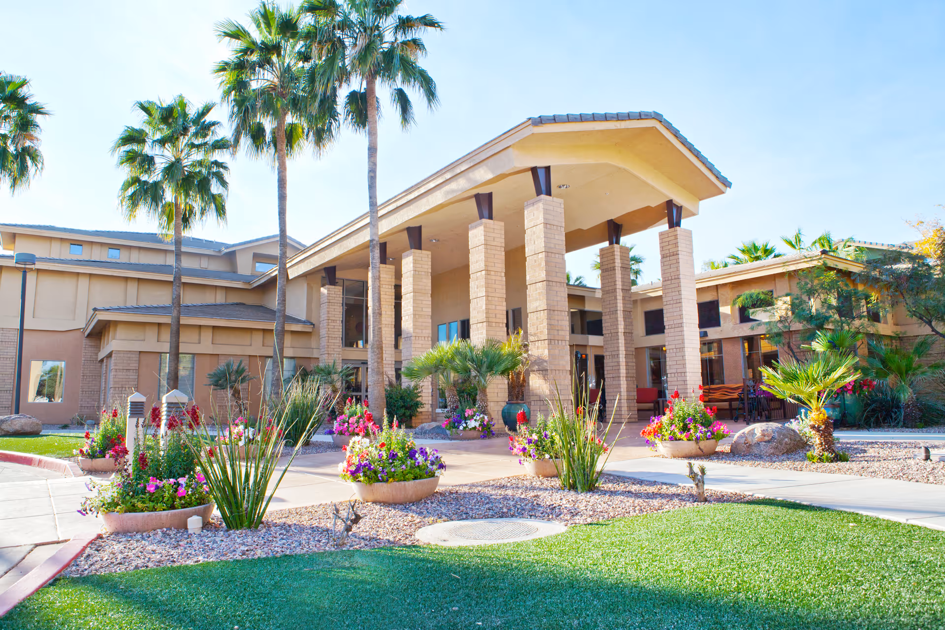 Exterior view of McDowell Village senior living facility showing a building with a covered entrance supported by brick columns, surrounded by palm trees, colorful flower beds, and well-maintained landscaping under a clear blue sky.