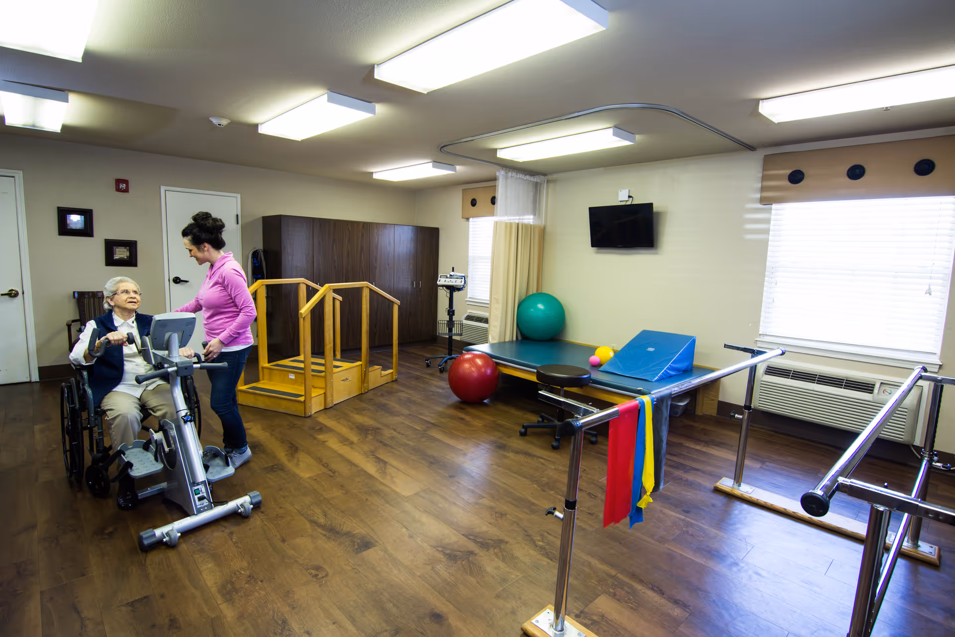 A physical therapy room in a nursing and rehab facility with wooden flooring, parallel bars, exercise balls, a small set of stairs with handrails, and a therapy table. An elderly woman in a wheelchair is using a stationary exercise bike with assistance from a female staff member wearing a pink top.