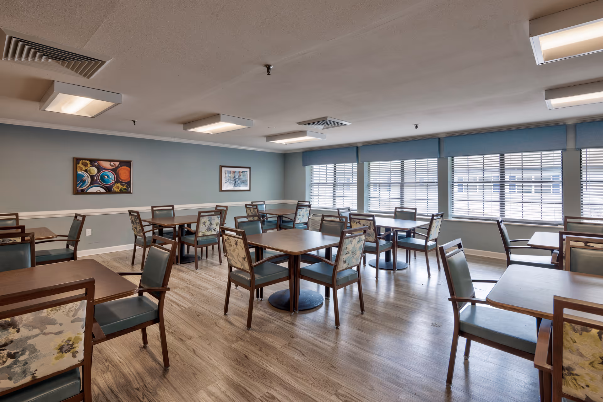 A dining room with multiple wooden tables and chairs arranged neatly on a wooden floor. The walls are painted light blue with white trim, and there are two framed pictures hanging on the wall. Large windows with blinds allow natural light to fill the room, and ceiling lights provide additional illumination.