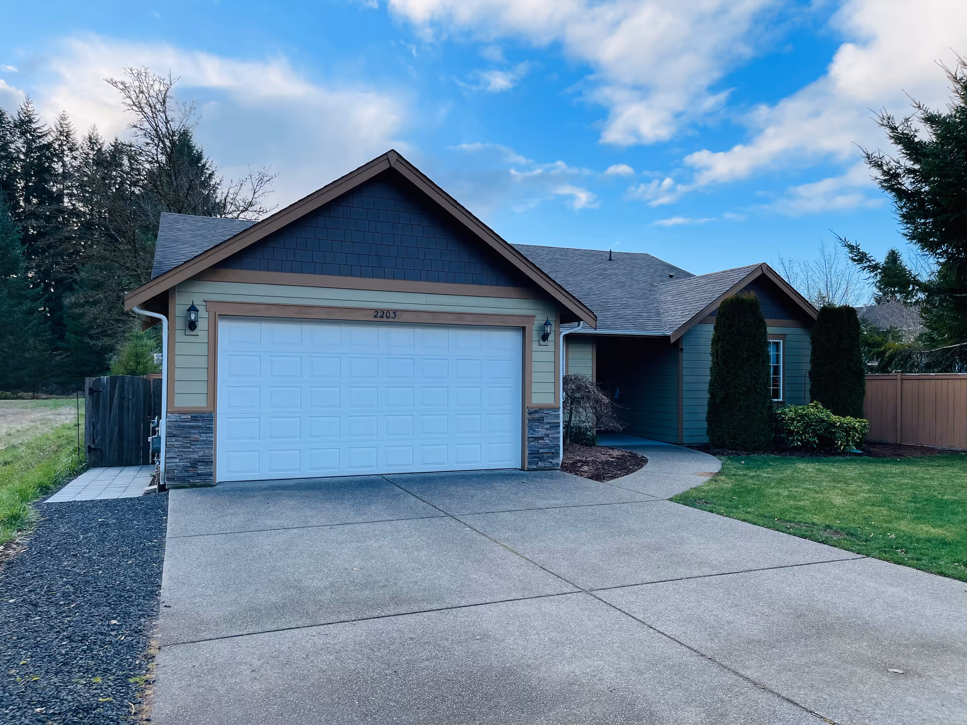 Single-story house with an attached two-car garage, driveway, front lawn, and trees under a partly cloudy sky.