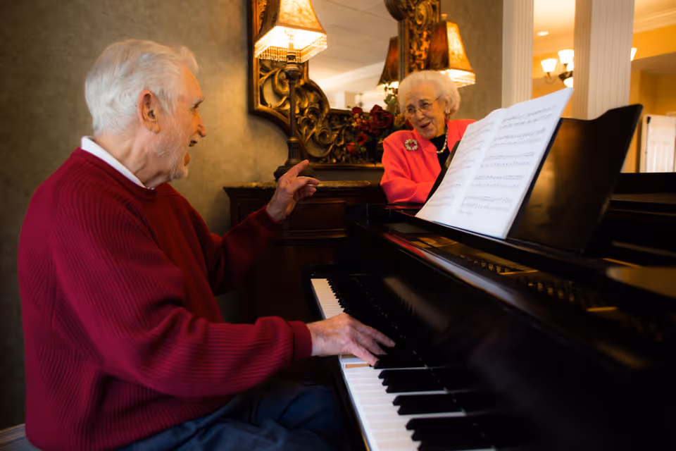An elderly man in a red sweater plays the piano while an elderly woman in a pink jacket watches and smiles. They are in a warmly lit room with ornate decor, including a large mirror and table lamps.