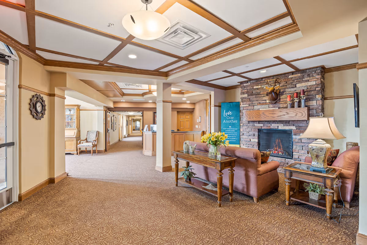 A cozy senior living facility common area with a stone fireplace, a mounted deer head above the mantel, and a lit fire. There are two upholstered armchairs and a sofa arranged around the fireplace, with wooden side tables holding a lamp and a vase of yellow flowers. The ceiling features wooden beams and recessed lighting. A hallway extends into the background with framed pictures on the walls and a wooden door at the end. A decorative clock is mounted on the left wall near the entrance.