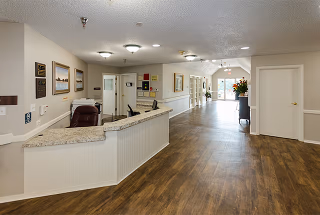 Interior view of a senior living facility reception area with a curved granite countertop desk, office chair, framed pictures on the wall, and a long hallway with wooden flooring leading to glass doors at the far end.