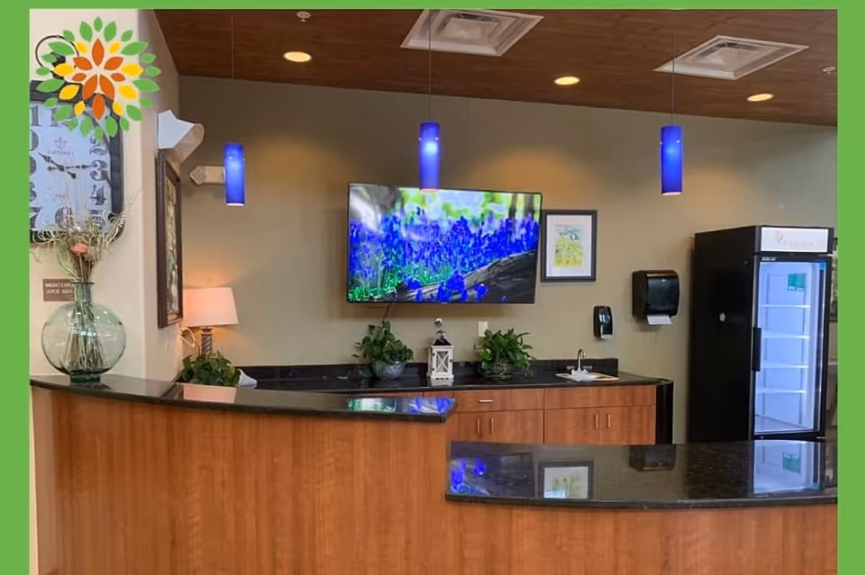 Reception area with a curved wooden counter topped with black granite. Behind the counter is a wall-mounted TV displaying a nature scene with blue flowers, two green plants, a lantern, and a paper towel dispenser. Three blue pendant lights hang from the ceiling. To the right is a black refrigerator with a glass door. On the left side, there is a large clock and a vase with dried flowers on the counter.