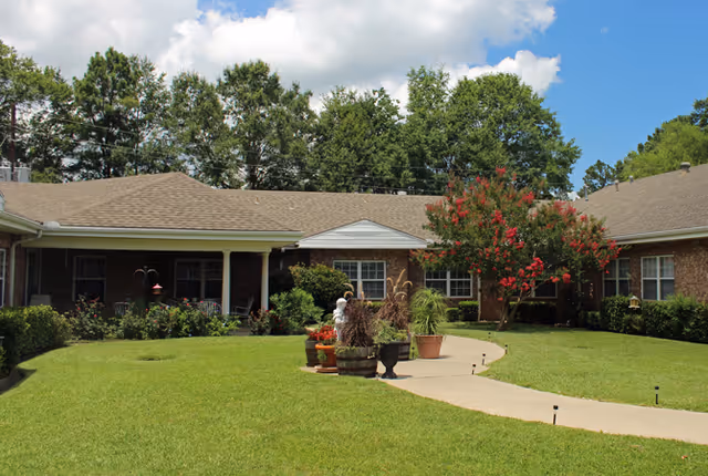 Single-story brick building with a curved walkway, manicured lawn, potted plants, and trees in a sunny courtyard.