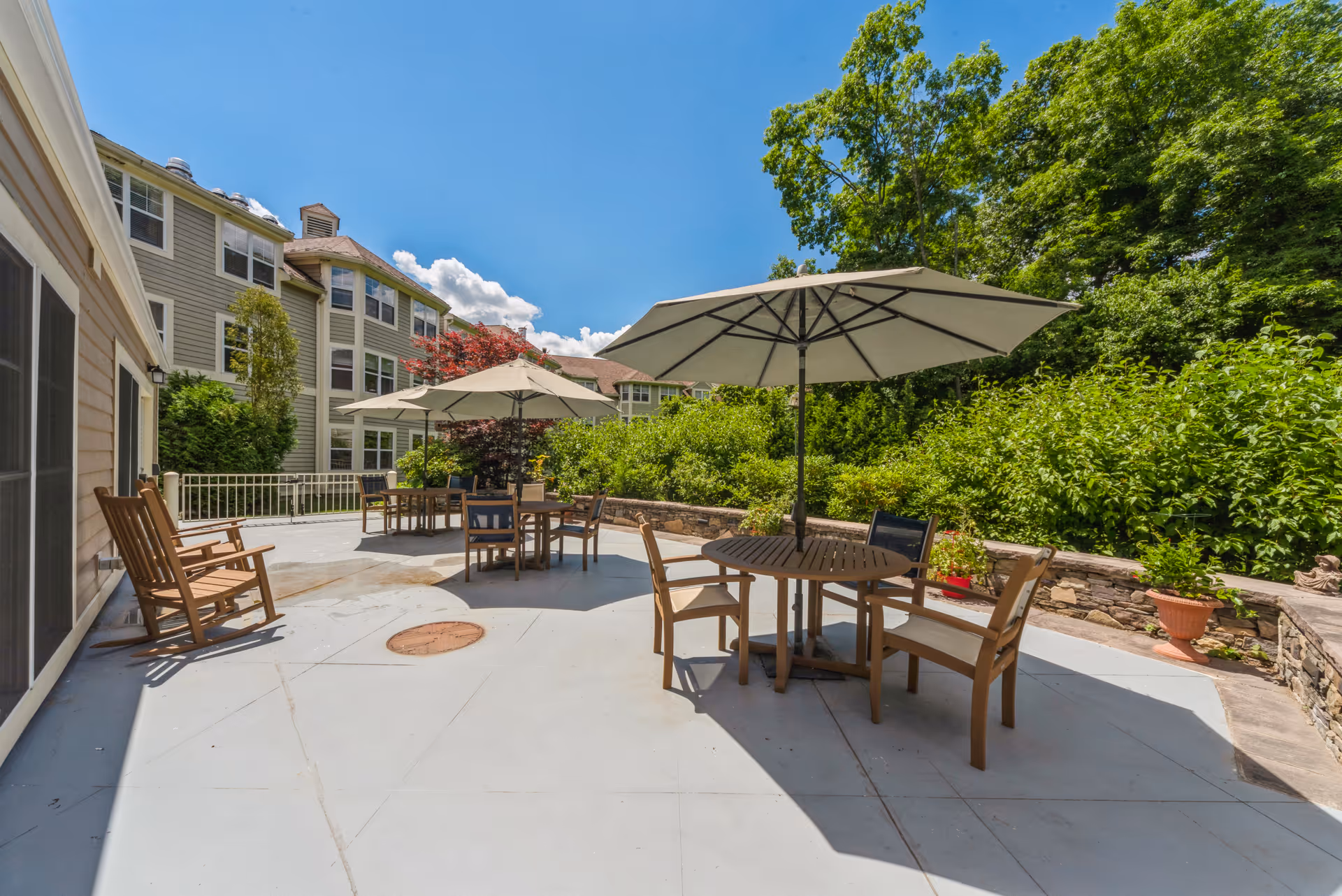 Sunny outdoor patio with round tables, large umbrellas, wooden chairs and rocking chairs beside a multi-story building and lush greenery.