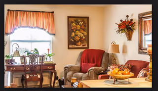 Cozy living room with upholstered armchairs, a wooden desk by a window, wall art, and a table topped with a bowl of fruit.