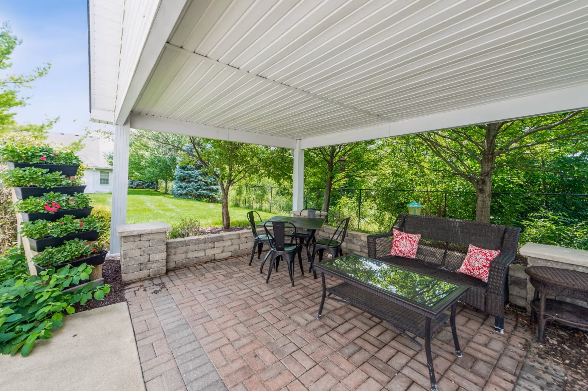 Covered outdoor patio area with brick flooring, a black metal table with four matching chairs, a black wicker loveseat with two red patterned cushions, a glass-top coffee table, and a tiered planter with green plants and flowers. The patio is surrounded by green trees and a grassy yard.