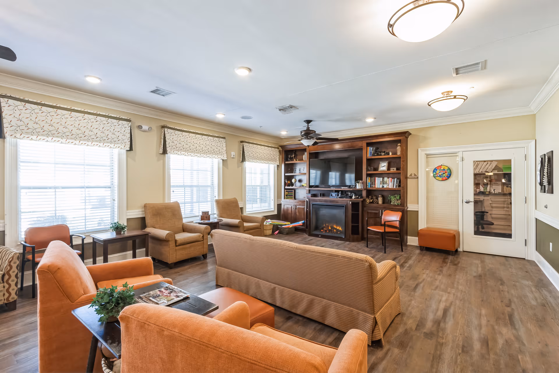 A cozy living room with multiple armchairs and a sofa arranged around a wooden entertainment center with a large flat-screen TV and an electric fireplace. The room has three large windows with patterned valances, wood flooring, and ceiling lights. There is a small table with a plant and magazines in the foreground and a door with glass panels on the right side.