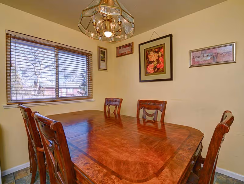 A dining room with a large wooden table and six matching wooden chairs. The room has light yellow walls with three framed pictures hanging, including one with flowers and another with the phrase 'Live Laugh Love'. A window with wooden blinds lets in natural light, and a brass chandelier with glass panels hangs above the table.