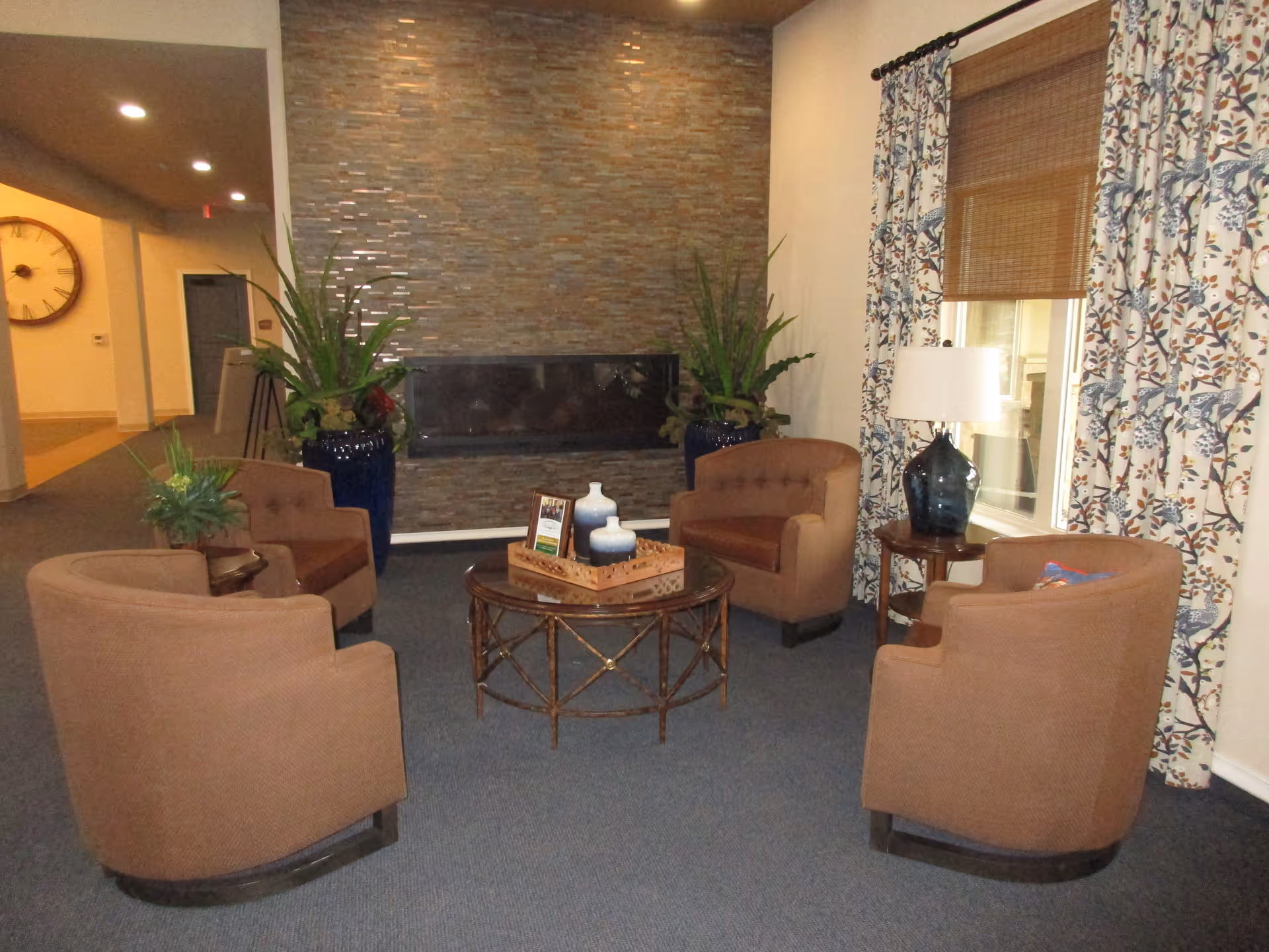 Cozy seating area with four brown armchairs arranged around a round glass-top coffee table in front of a tiled fireplace wall, with plants and a lamp by a curtained window.