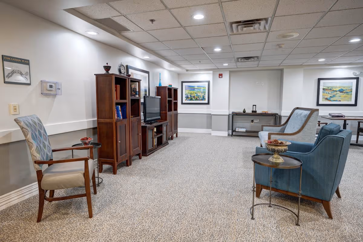 A well-lit common area with carpeted floor featuring wooden bookshelves, a television, framed artwork on the walls, and several upholstered chairs arranged around small tables with decorative bowls.