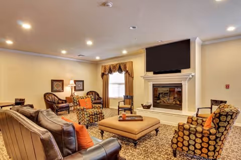 Comfortable senior living lounge with sofas, armchairs, an ottoman, fireplace, and wall-mounted TV.