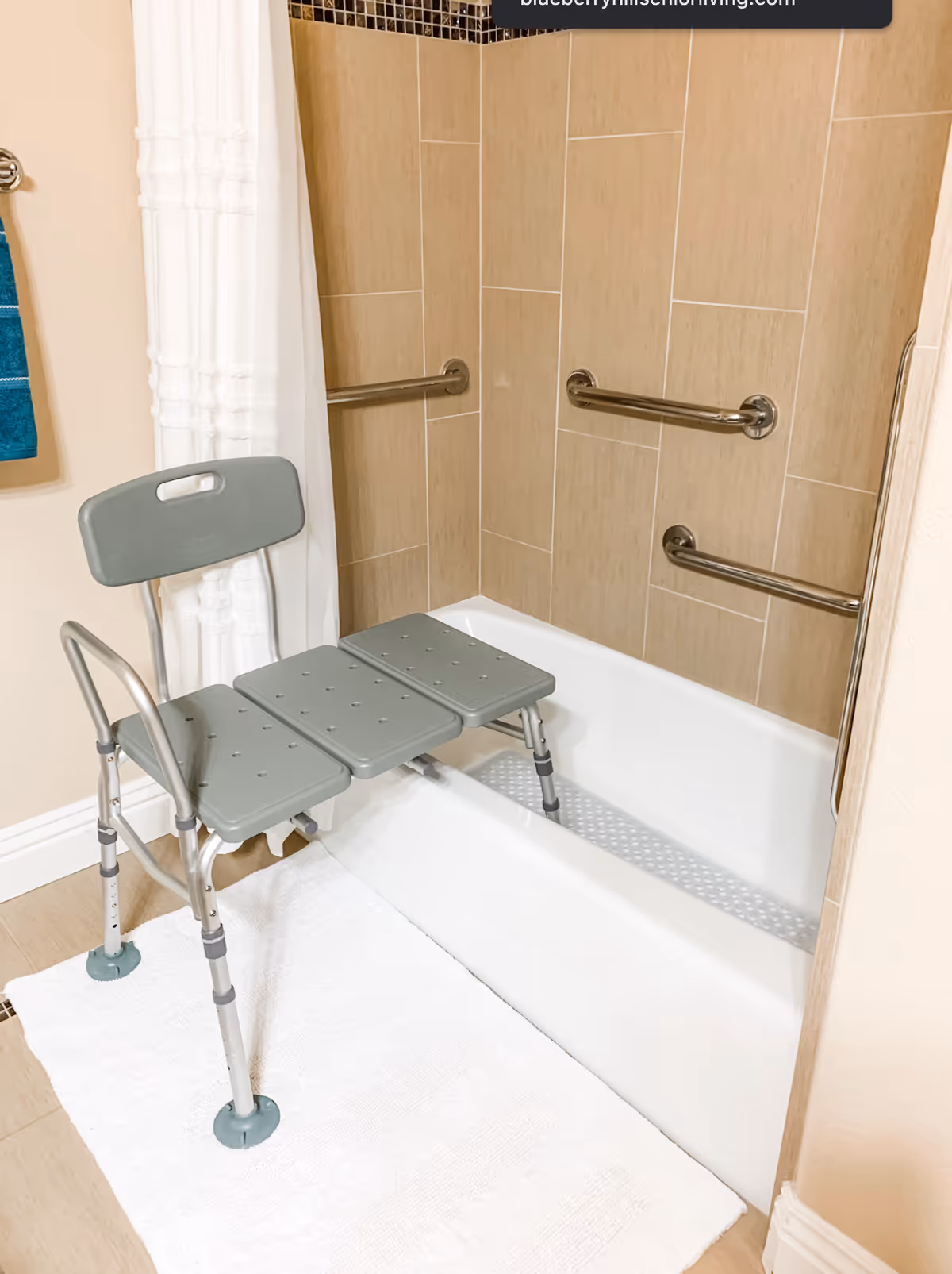 A bathtub with beige tiled walls and three metal grab bars installed inside. A gray shower chair with a backrest is placed partially inside the tub, and a white bath mat is on the floor in front of the tub. A white shower curtain and a blue towel hanging on a wall-mounted towel rack are also visible.