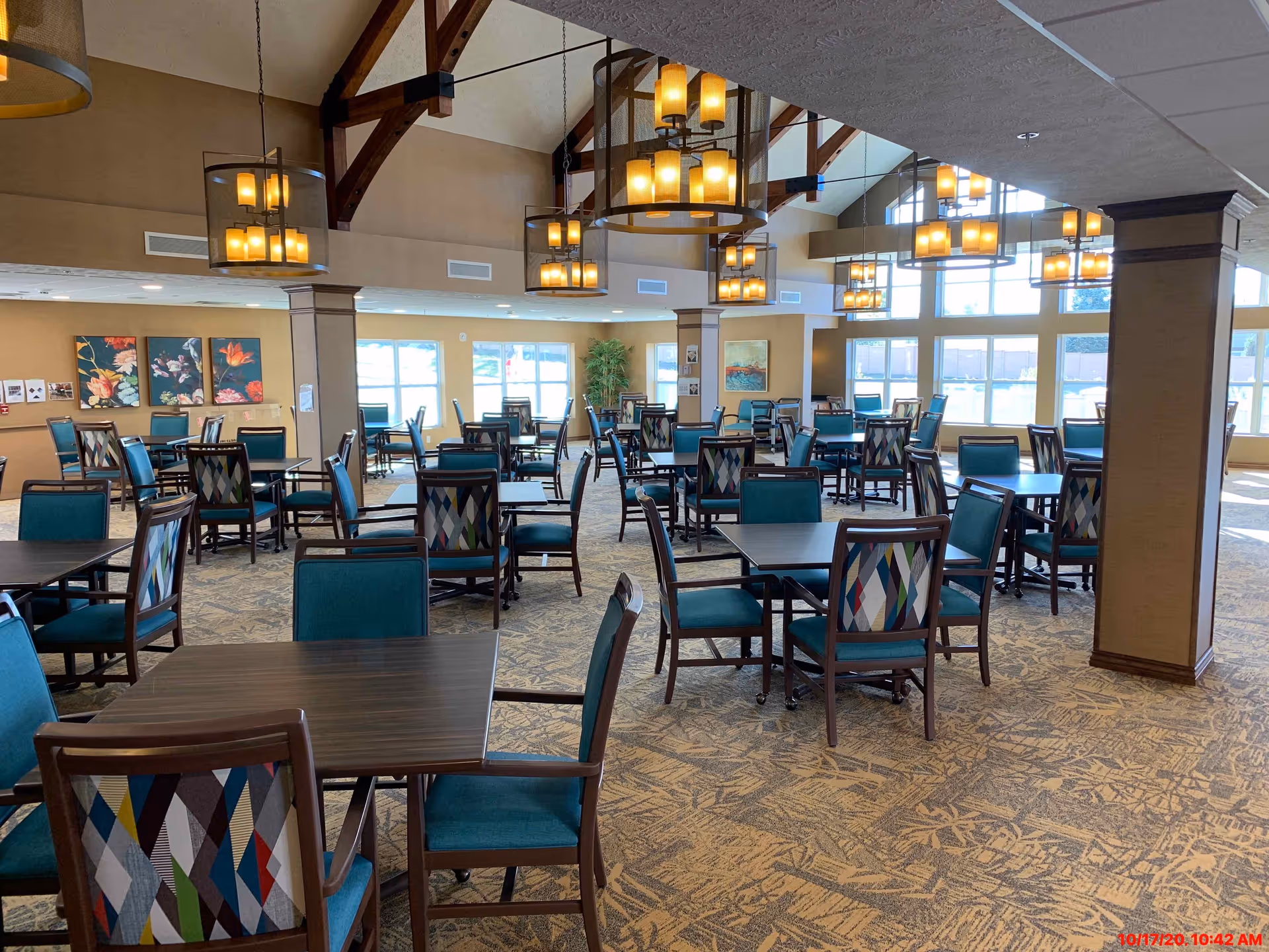 A spacious dining room with multiple dark wooden tables and chairs featuring teal cushions and multicolored patterned backs. The room has large windows allowing natural light to fill the space, beige walls, carpeted floor with a patterned design, and several hanging chandeliers with warm lighting. There are decorative paintings on the walls and wooden beams supporting the ceiling.