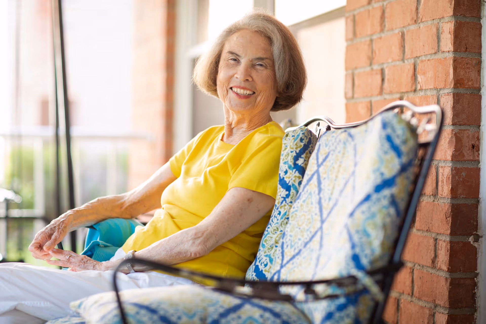 An elderly woman with short gray hair wearing a yellow shirt and white pants is sitting and smiling on a cushioned outdoor bench with a brick wall in the background.