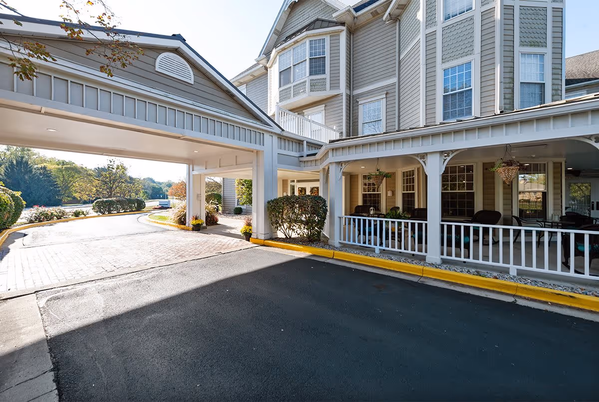 Exterior view of a senior living facility named Bloom at Kessler showing a covered driveway entrance with a paved road, landscaped bushes, and a porch area with seating and hanging plants.