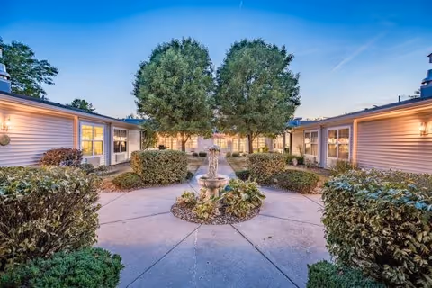 Outdoor courtyard area at dusk with a central fountain surrounded by bushes and trees, flanked by single-story buildings with lit windows.