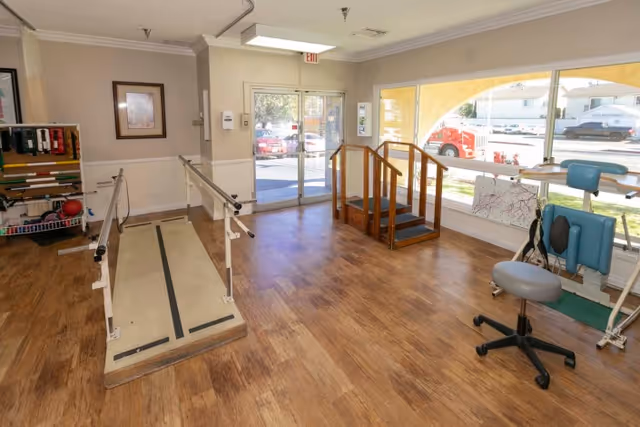 Bright rehabilitation room with parallel walking bars, wooden practice steps, therapy chairs and equipment near a glass entrance and large window.