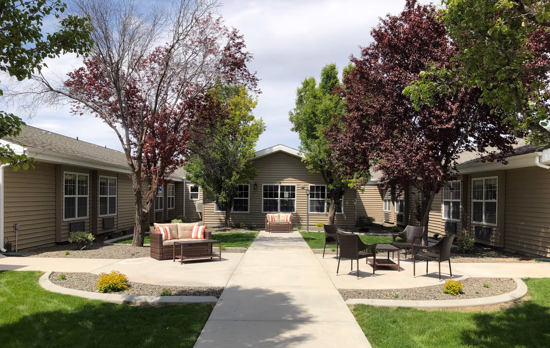 Outdoor courtyard area at The Gables Assisted Living of Caldwell featuring a concrete walkway, green grass, several trees with red and green leaves, and outdoor seating including cushioned sofas and chairs with small tables.
