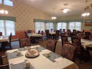 A dining room with multiple tables covered with white tablecloths and place settings including cups and napkins. The room has several windows with blinds and valances, patterned wallpaper, and ceiling fans with lights. Chairs with floral upholstery surround the tables.
